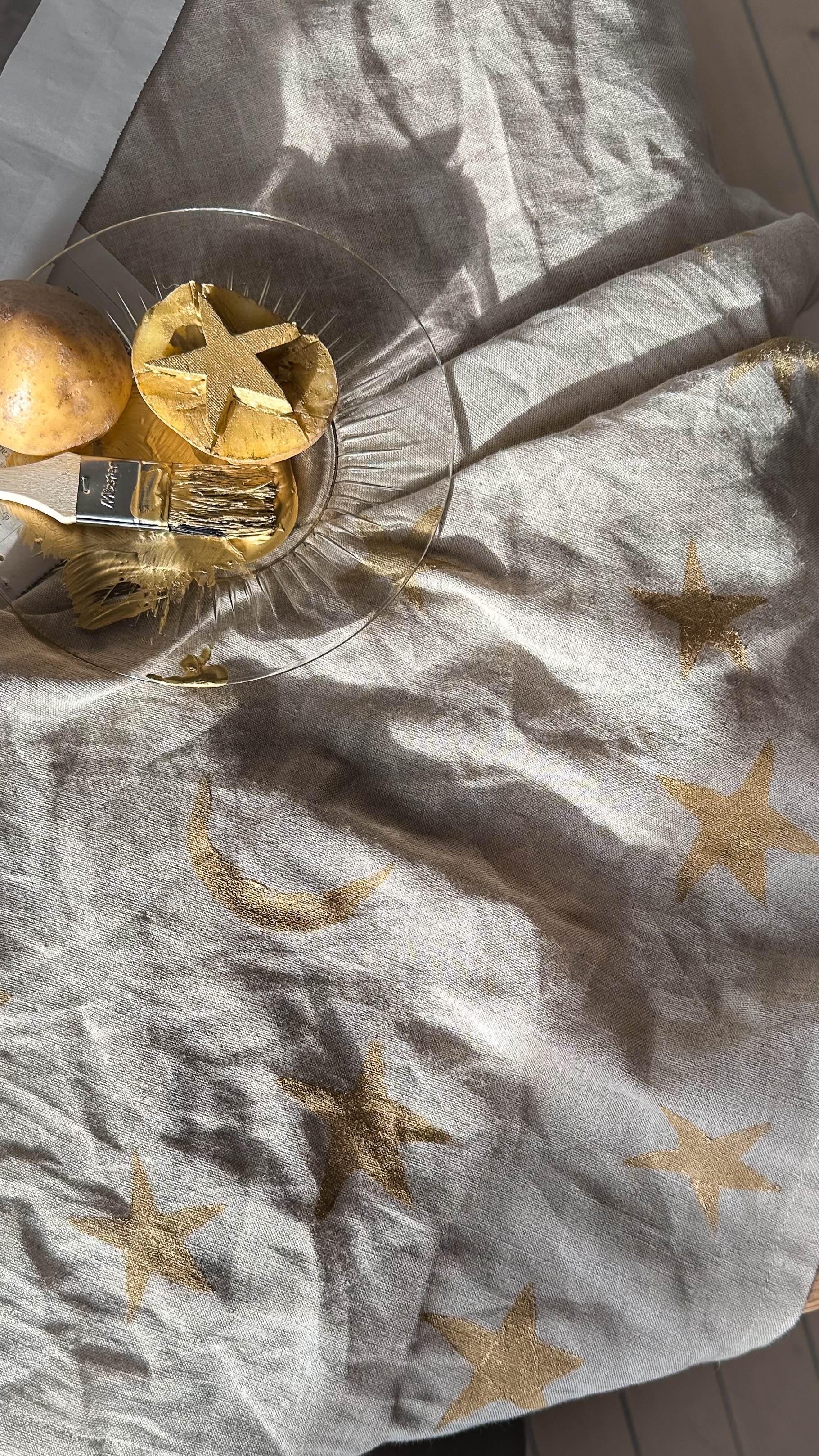 A star-cut potato, paintbrush with gold paint, and light fabric adorned with gold stars and a crescent moon.