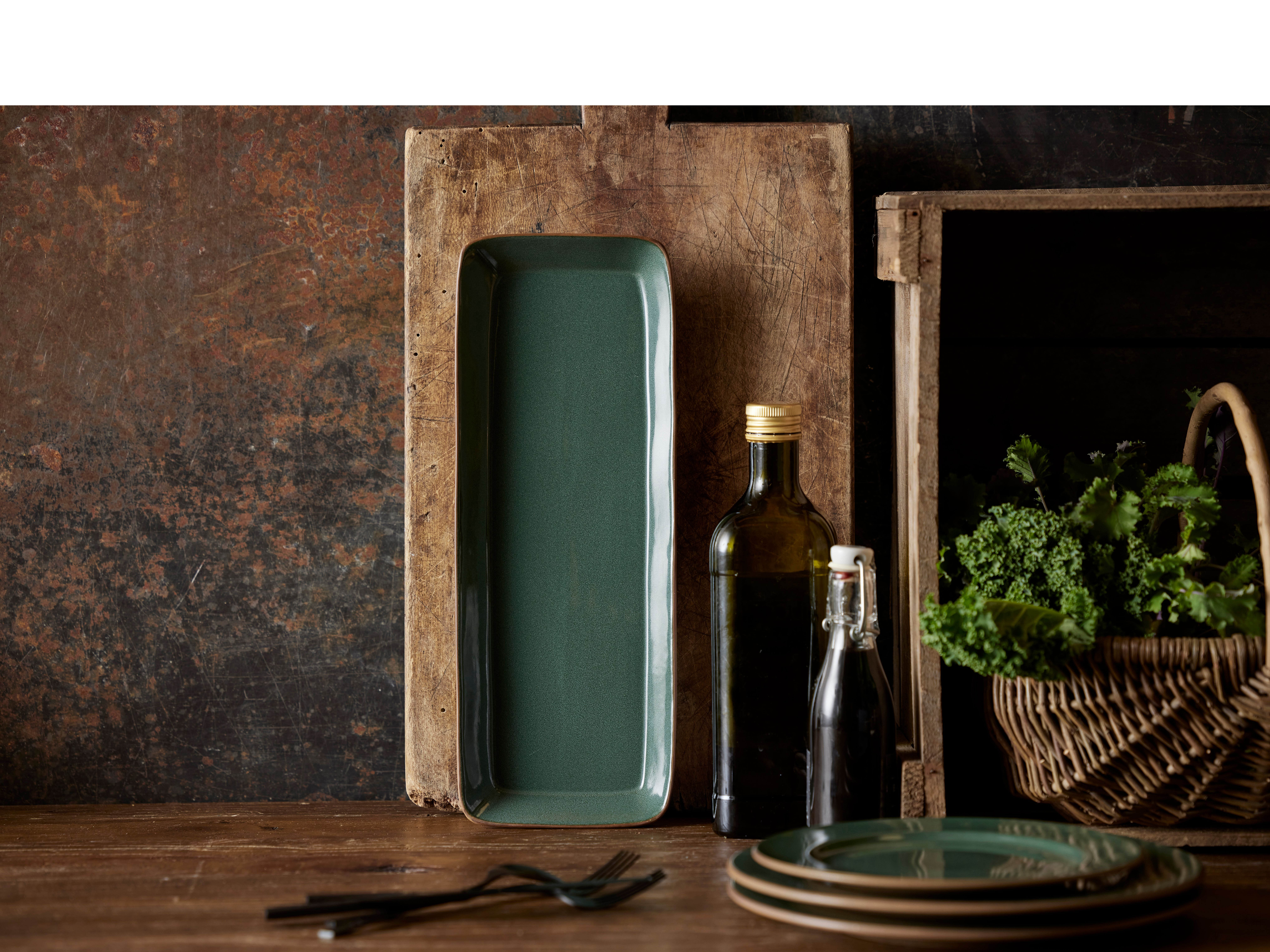 ett träbord med tallrikar, bestick, en flaska olja och en korg med grönsaker.