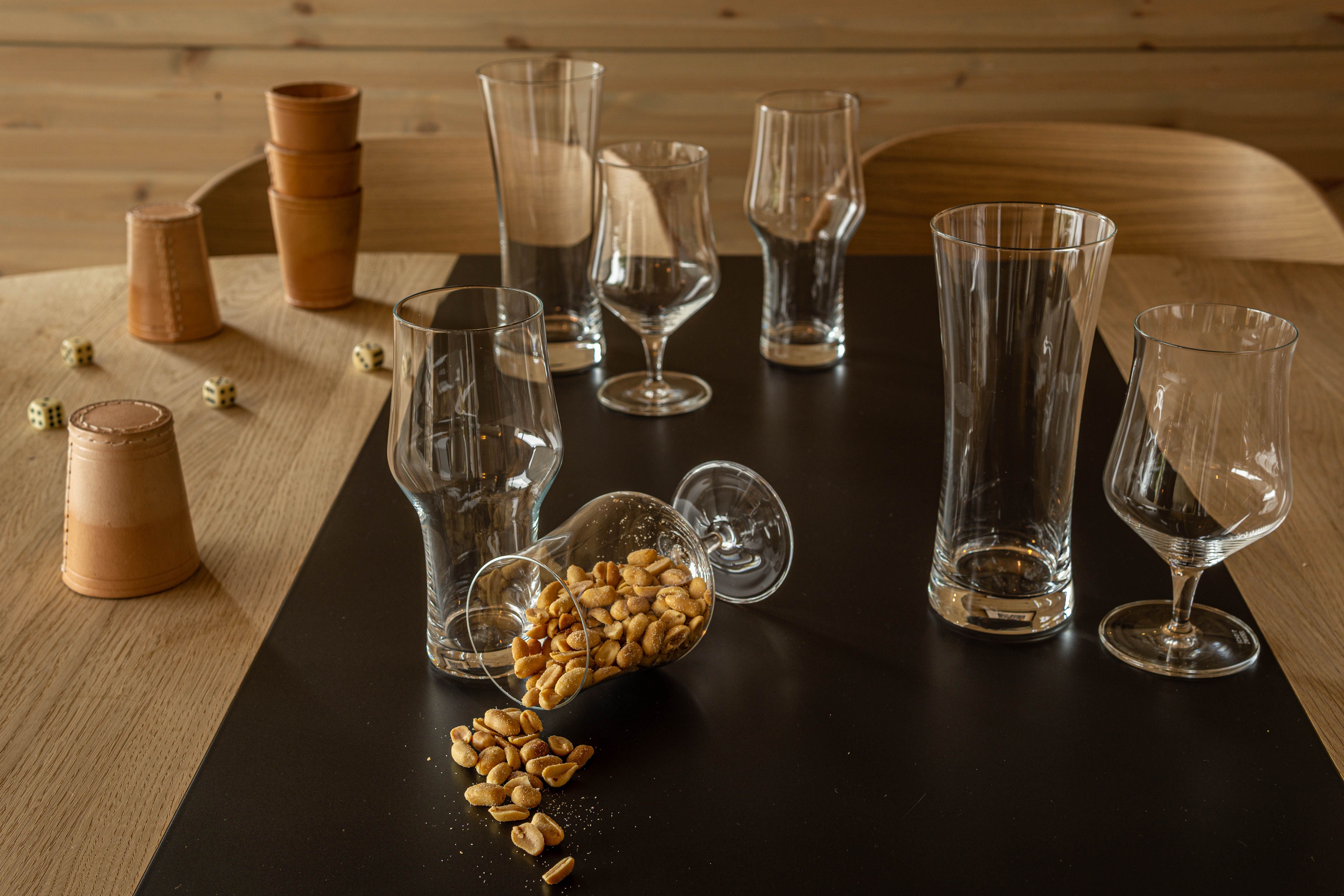 Tomma ölglas och vinglas står på ett bord med ett omkullvält glas fyllt av jordnötter samt tärningar och lädermuggar i bakgrunden.