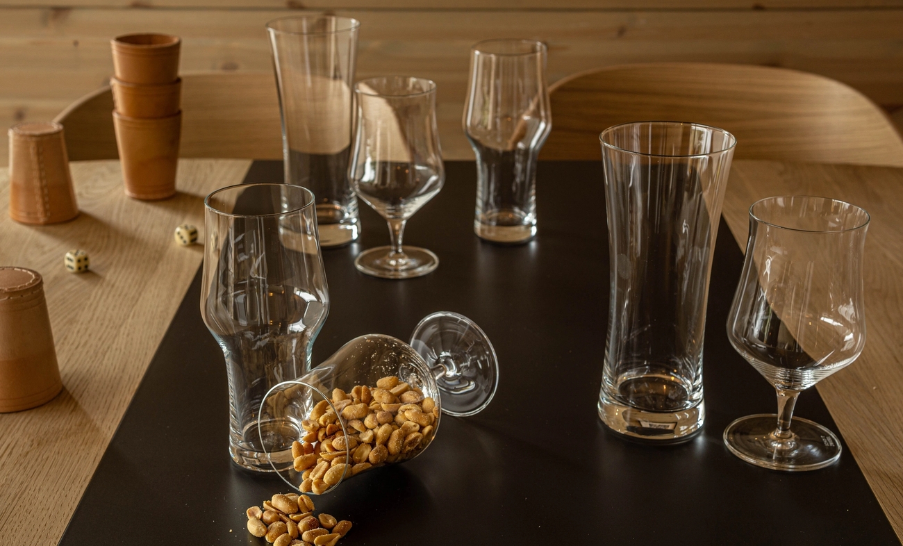 Tomma ölglas och vinglas står på ett bord med ett omkullvält glas fyllt av jordnötter samt tärningar och lädermuggar i bakgrunden.