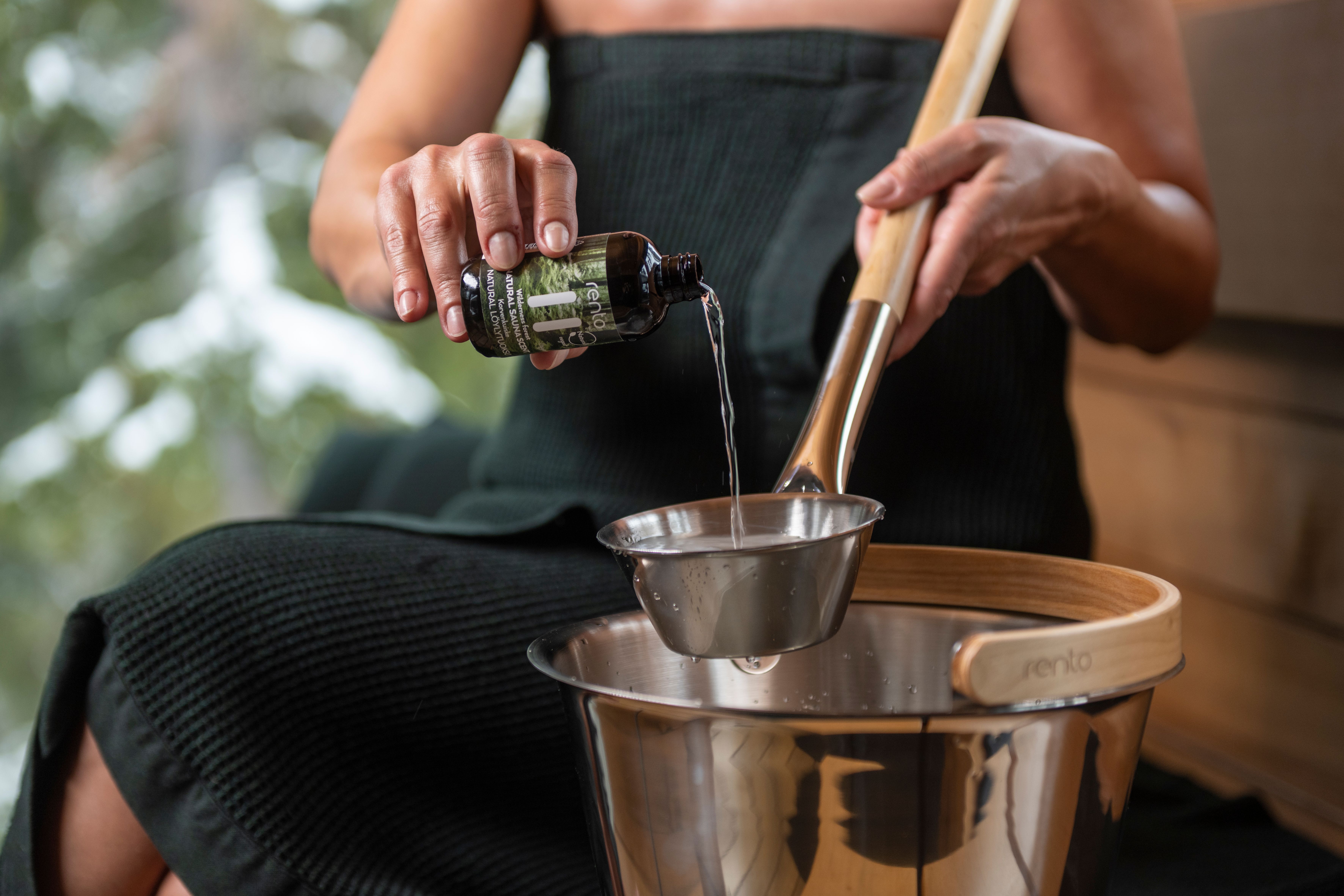 Henkilö kaataa nestettä pullosta kauhaan saunaämpärin yläpuolella, ulkona lunta.