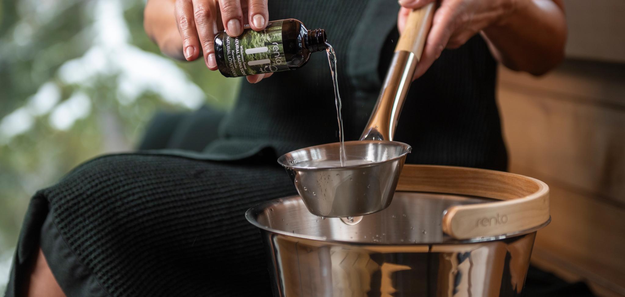 Henkilö kaataa nestettä pullosta kauhaan saunaämpärin yläpuolella, ulkona lunta.