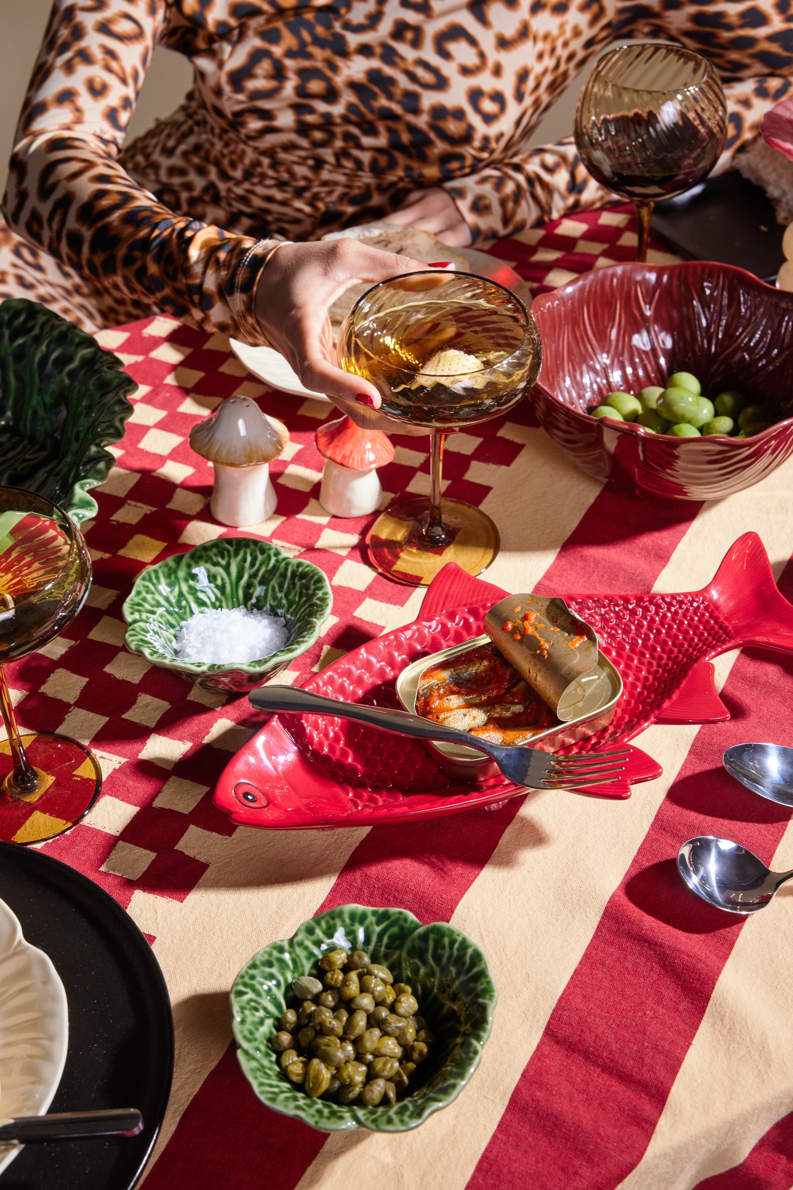 Eclectic table setting on a red and beige checkered cloth, with a person in leopard print, mushroom salt shaker, green leaf dishes, and a red fish dish.