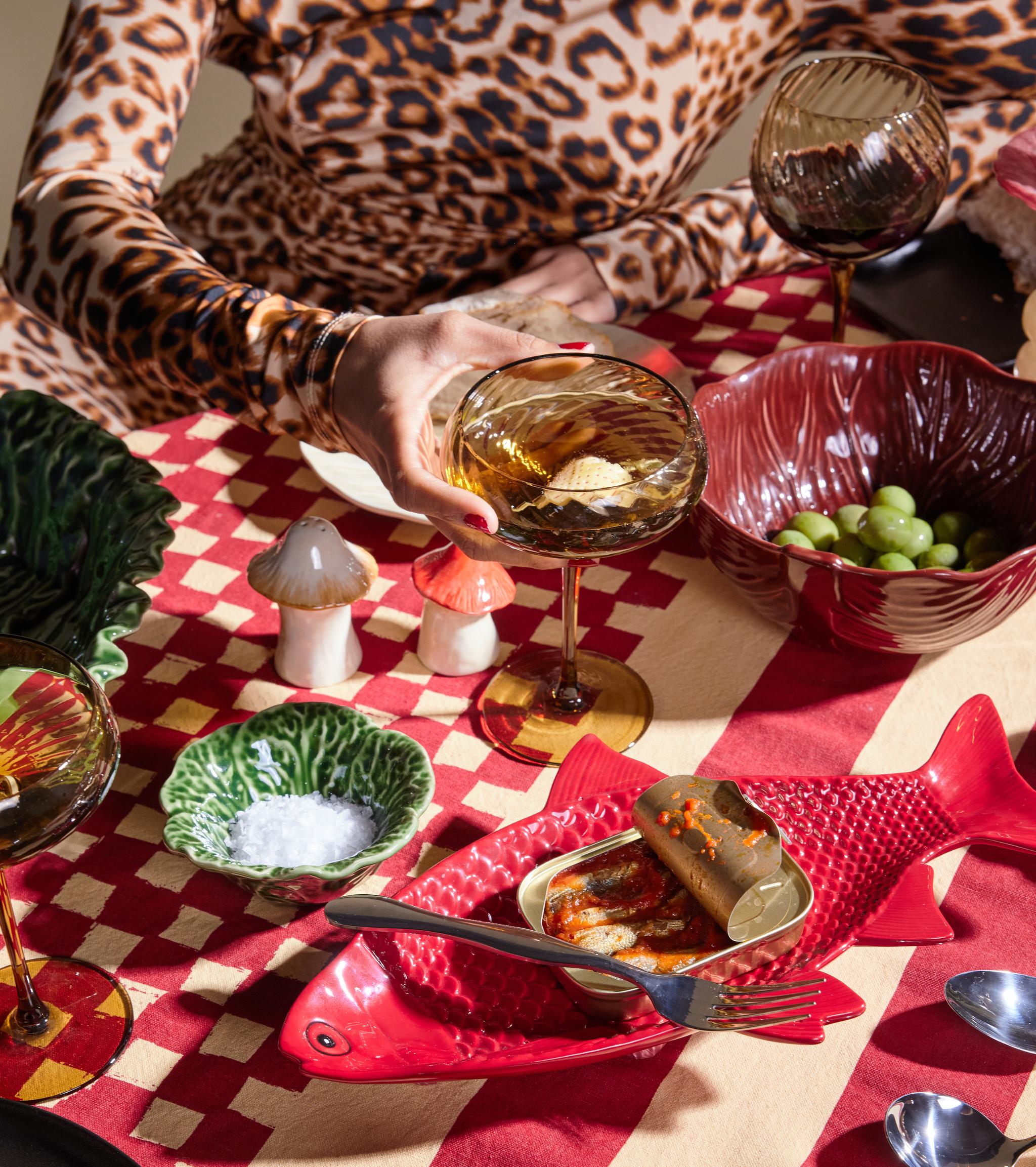 Eclectic table setting on a red and beige checkered cloth, with a person in leopard print, mushroom salt shaker, green leaf dishes, and a red fish dish.