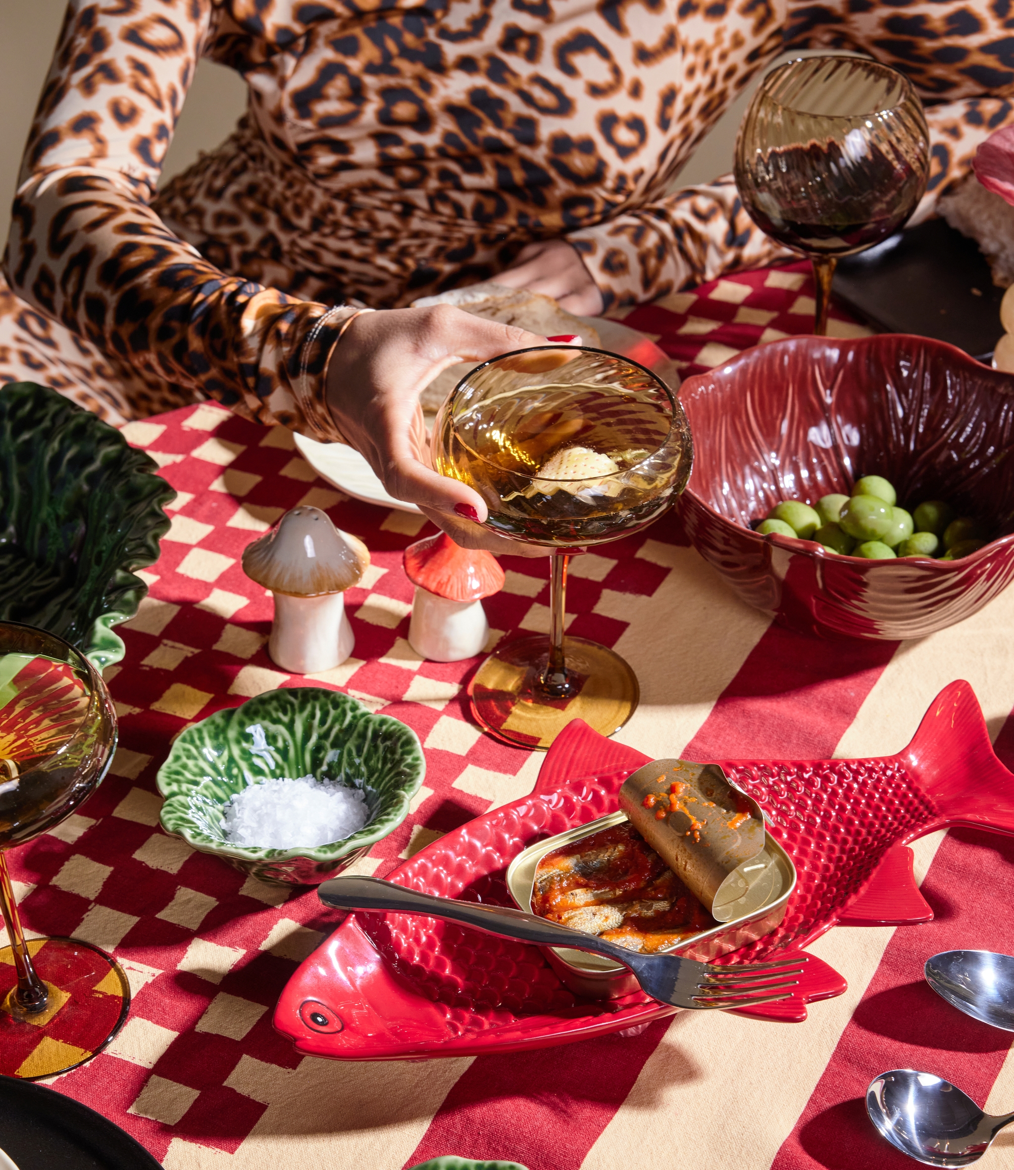 A person in a leopard print top holds a drink at a table set with a red and cream checkered tablecloth, featuring an open sardine tin on a red fish plate, bowls of salt, capers, and olives, and mushroom shakers.