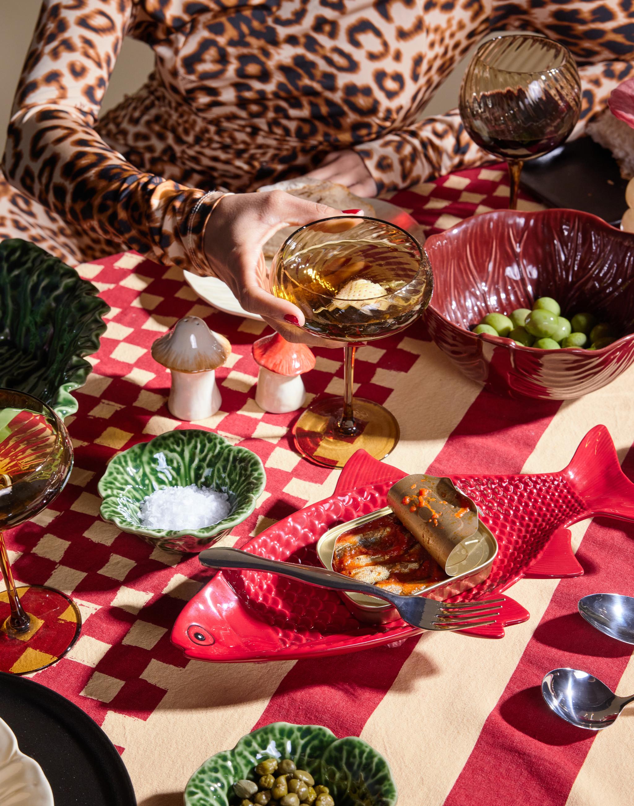 En person i leopardmønster holder en drink ved et bord dekket med en åpen sardinboks på en rød fiskeformet tallerken, skåler med oliven, kapers og salt, alt på en rød- og kremfarget mønstret duk