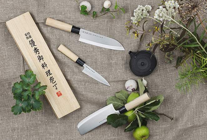 Japanilaiset keittiöveitset puisine kahvoineen aseteltuna yrttejen, vihannesten ja puisessa rasiassa olevan pakkauksen kanssa pellavakankaalle.