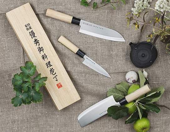 Japanilaiset keittiöveitset puisine kahvoineen aseteltuna yrttejen, vihannesten ja puisessa rasiassa olevan pakkauksen kanssa pellavakankaalle.