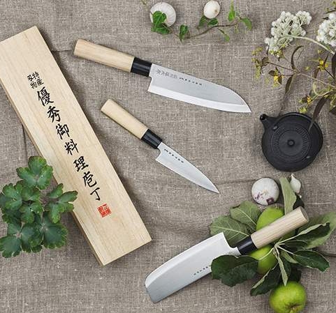 Japanilaiset keittiöveitset puisine kahvoineen aseteltuna yrttejen, vihannesten ja puisen lahjalaatikon kanssa pellavakankaalle.