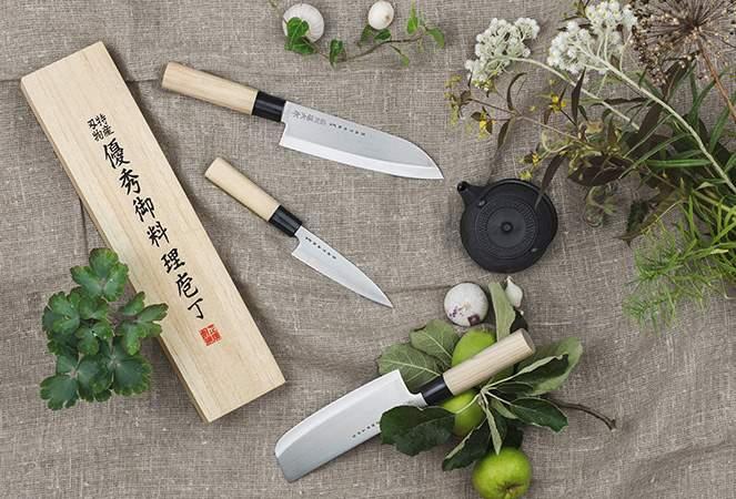 Japanilaiset keittiöveitset puisine kahvoineen aseteltuna luonnonkasvien, vihreiden omenoiden ja puisen lahjalaatikon kanssa pellavakankaalle.