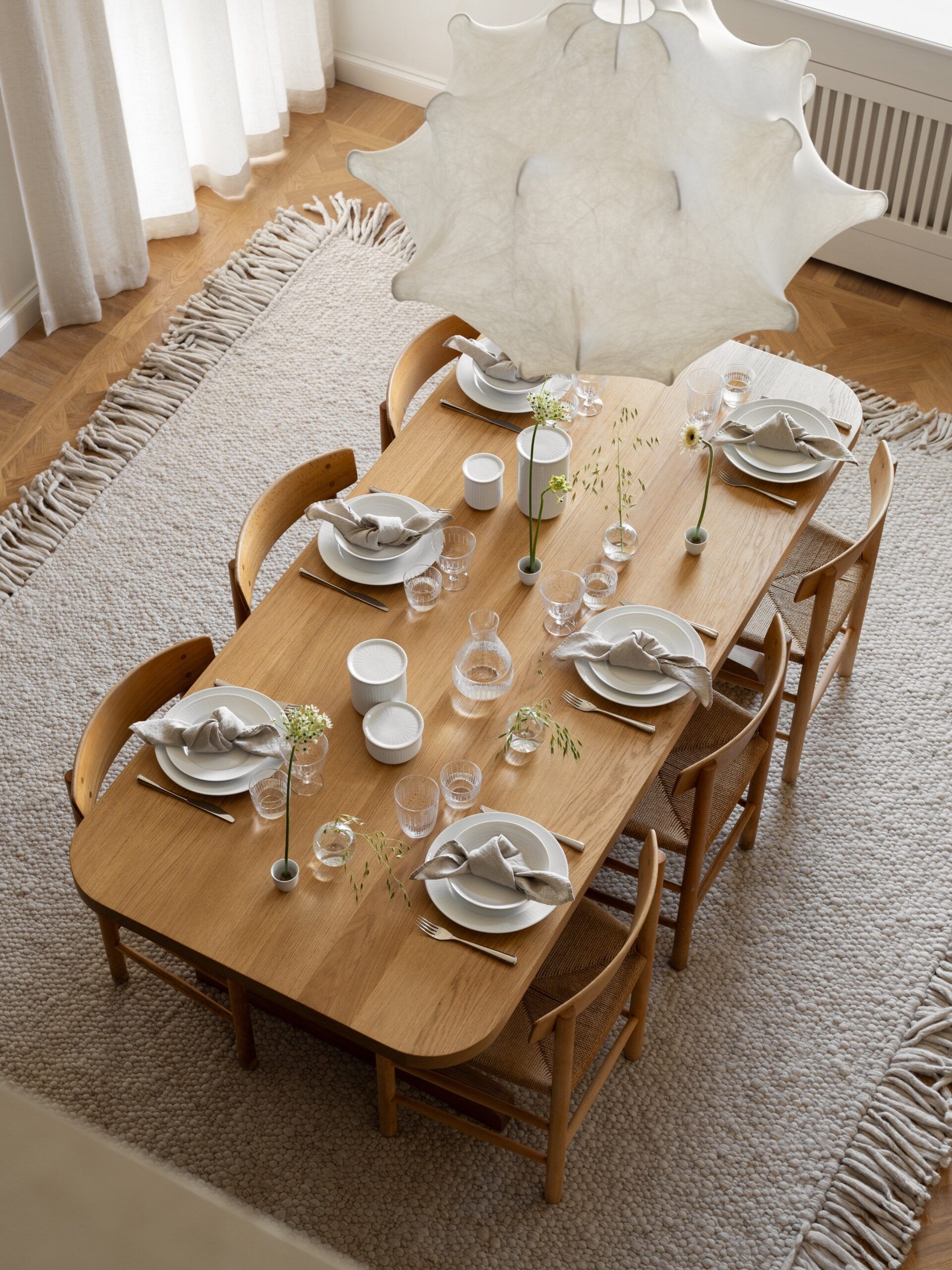 Luftfoto av et moderne spisebord i tre med hvite tallerkener og små blomster, på et teksturert teppe, under en stor hvit lysekrone.