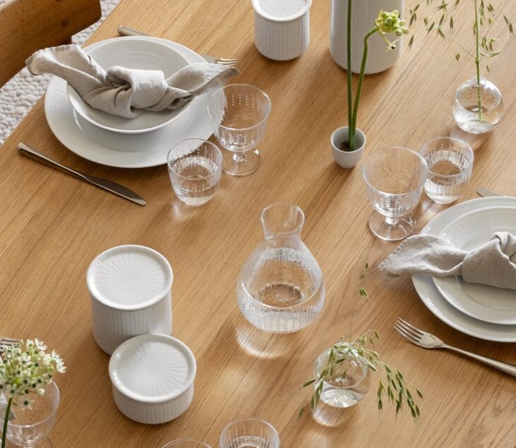 Ylhäältä katsottuna puinen ruokapöytä, jossa on lautasia, pellavaisia lautasliinoja, minimalistisia kukka-asetelmia ja laseja vaalealla teksturoidulla matolla.