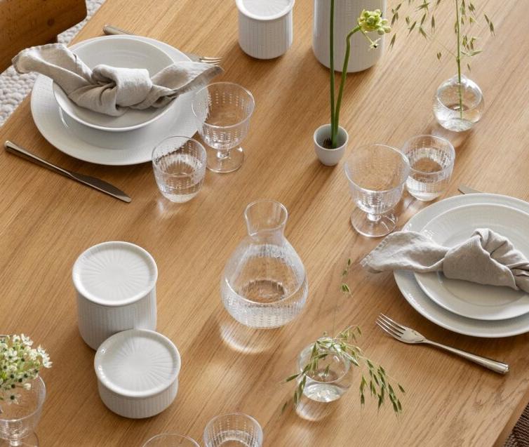 Ylhäältä katsottuna puinen ruokapöytä, jossa on lautasia, pellavaisia lautasliinoja, minimalistisia kukka-asetelmia ja laseja vaalealla teksturoidulla matolla.