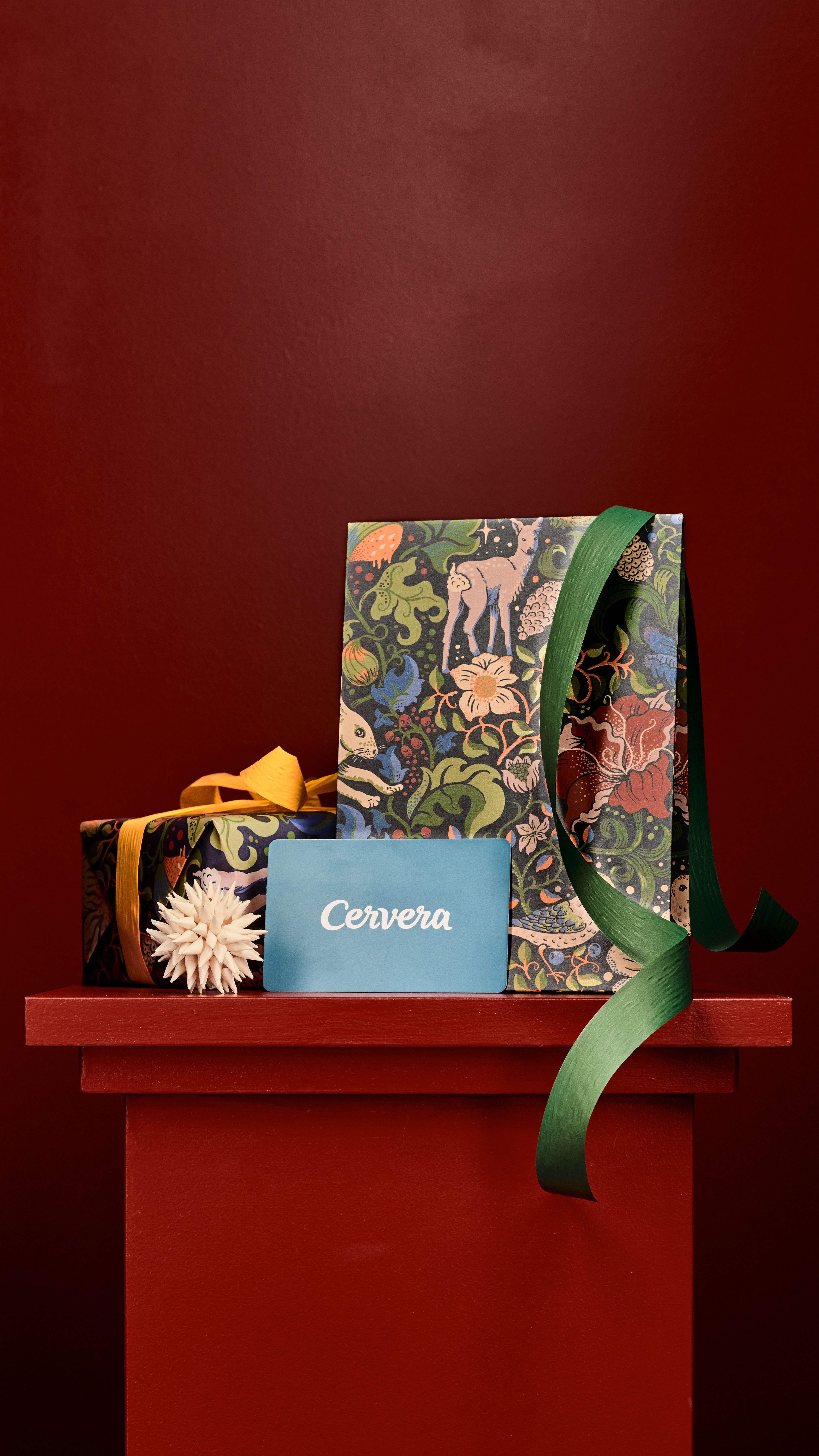 Två presenter inslagna i mörkt, naturmönstrat papper med ett blått "Cervera"-presentkort på en röd hylla.