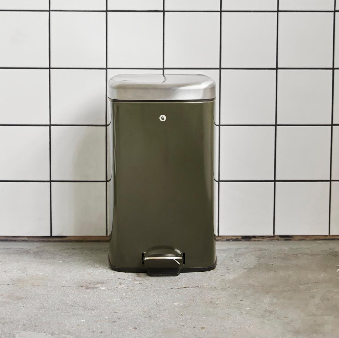Grön pedalhink i metall står mot en vit kakelvägg med svarta fogar på ett betonggolv.