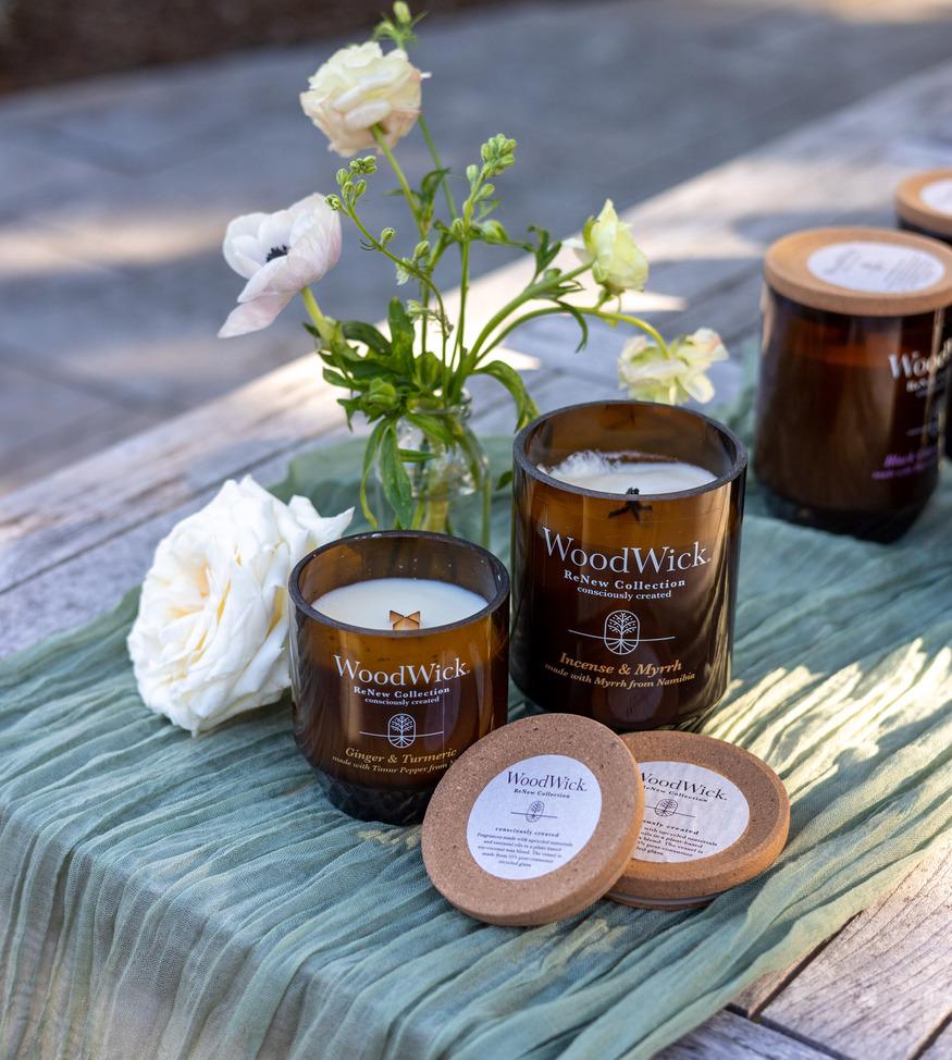 Tre ljus från WoodWick ReNew Collection i bärnstensfärgade glasburkar med korklock, arrangerade med vita blommor på en grön tyglöpare på ett träbord.