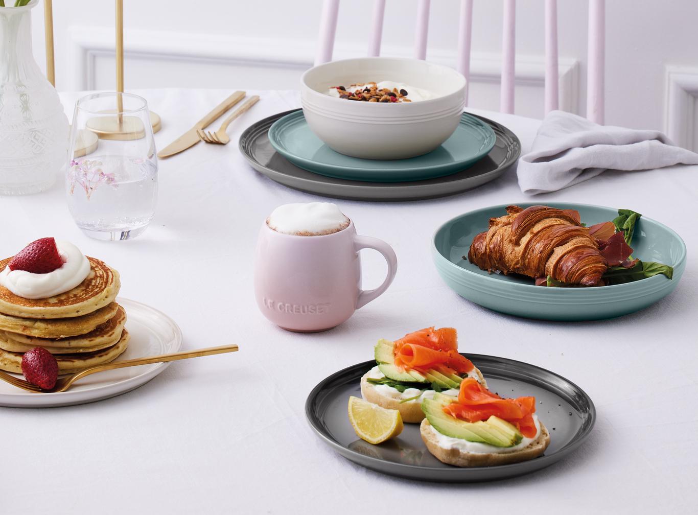 En ljus, pastellfärgad frukost serverad på ett vitt bord, med pannkakor, en croissant, yoghurt, kaffe, blommor och tända ljus.