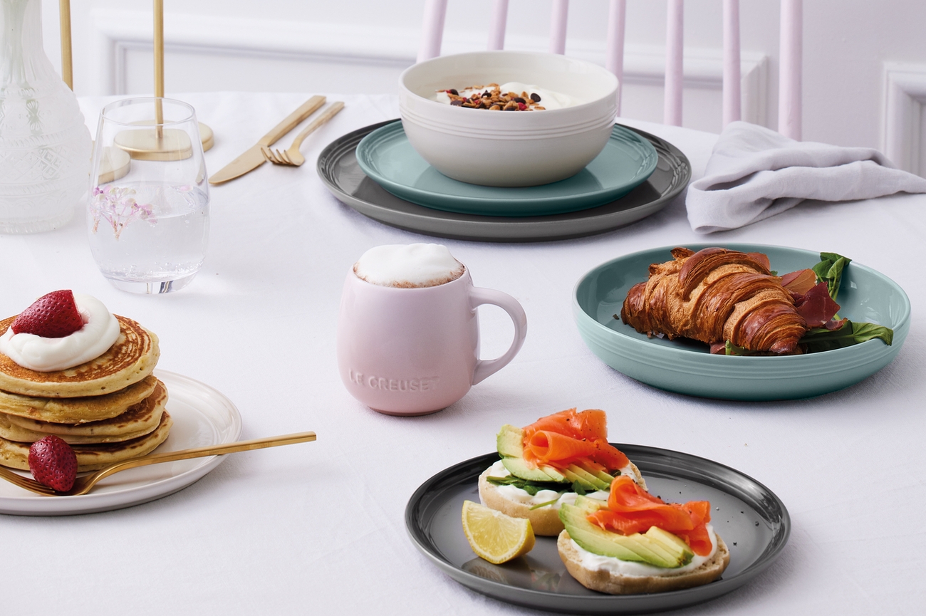 En ljus frukost med pannkakor med jordgubbar, bagels med lax och avokado, en croissant och yoghurt, serverad på pastell- och neutralfärgade fat.