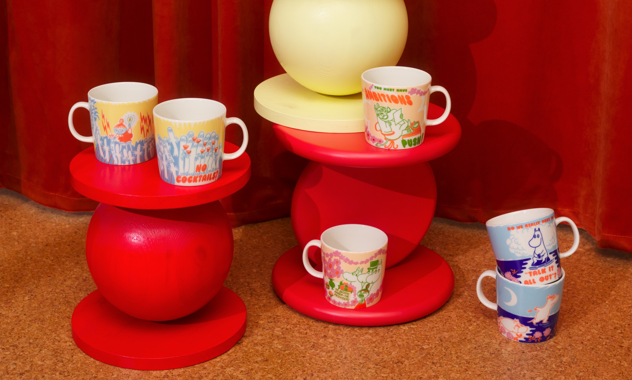 Värikkäät Muumi-mukit on aseteltu punaisille ja keltaisille design-jakkaroille lämpimän oranssin taustan eteen.