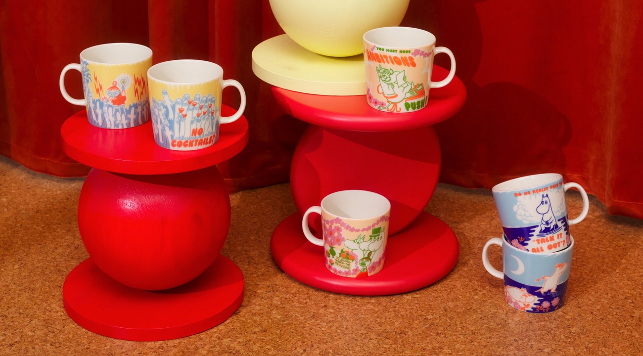 Värikkäät Muumi-mukit on aseteltu punaisille ja keltaisille design-jakkaroille lämpimän oranssin taustan eteen.