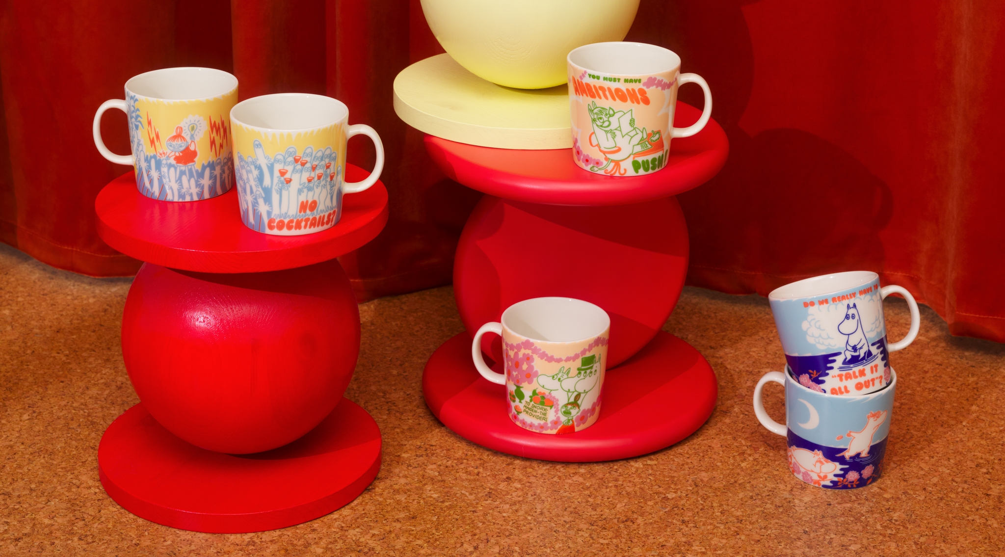 Värikkäät Muumi-mukit on aseteltu punaisille ja keltaisille design-jakkaroille lämpimän oranssin taustan eteen.