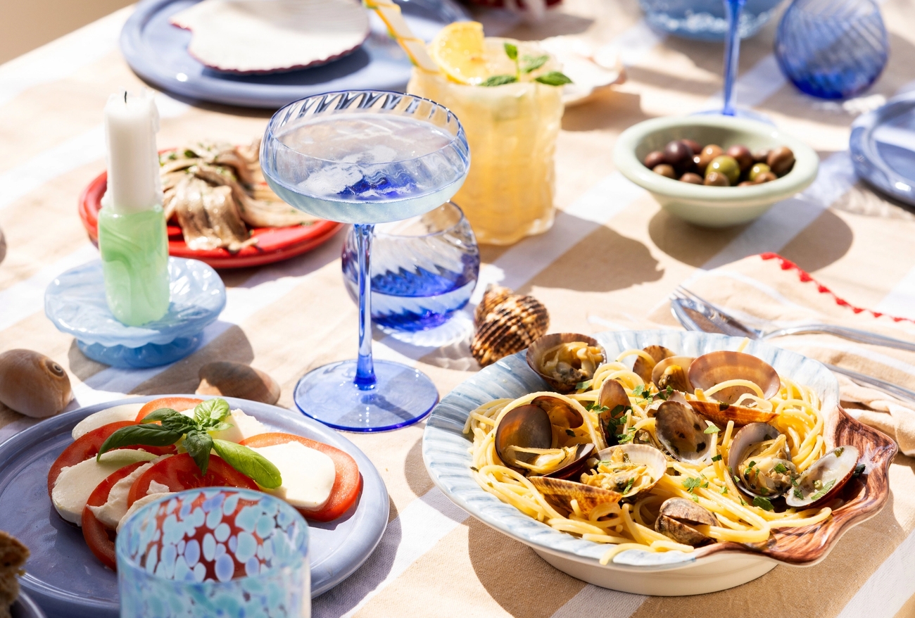 Somrig dukning med pasta, skaldjur, sallad och färgglada glas på ett bord vid havet.
