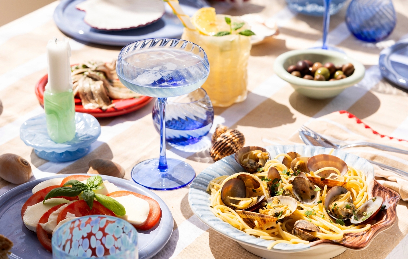 Somrig dukning med pasta, antipasti och färgglada glas på ett bord vid havet.