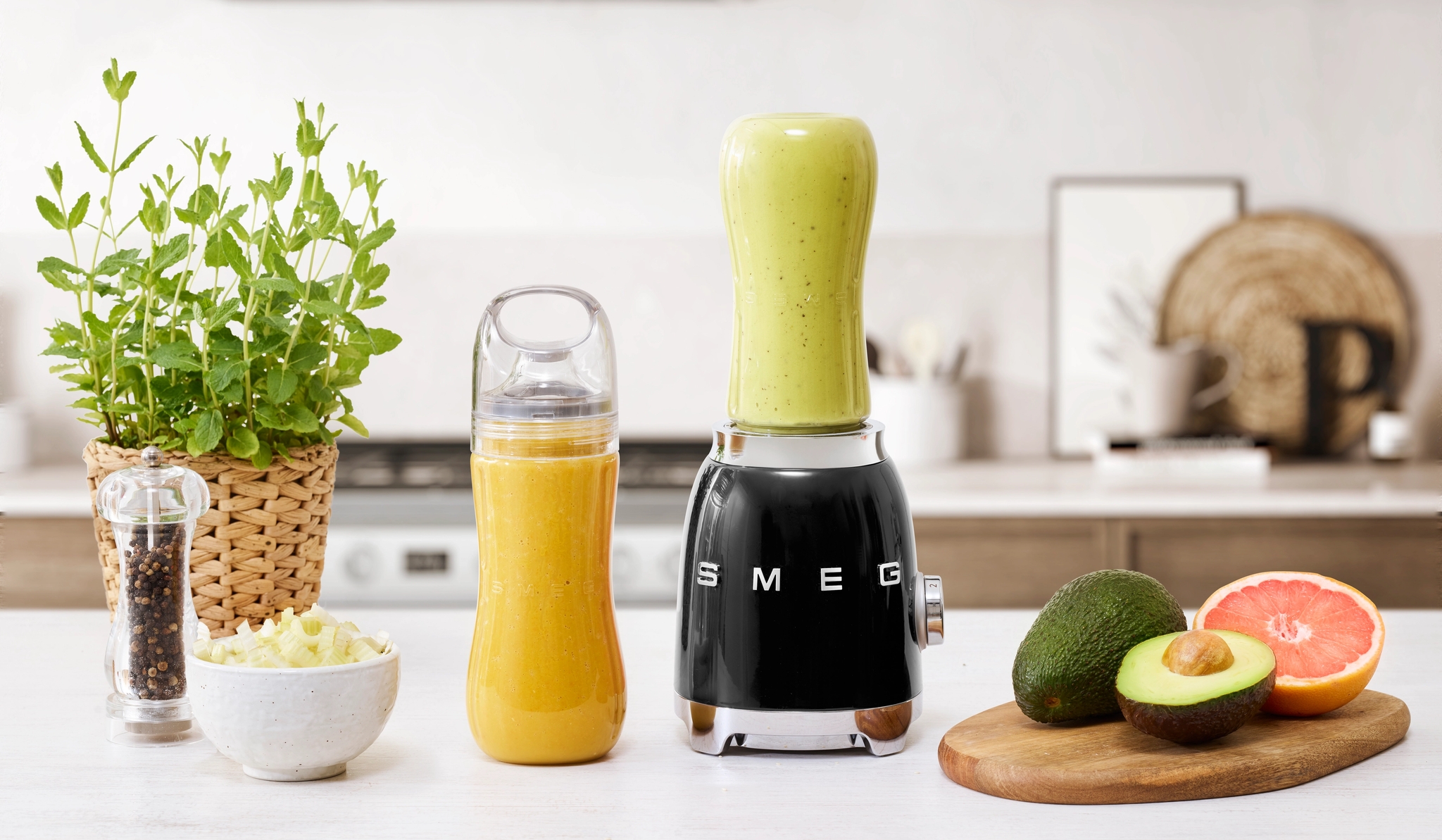 En svart Smeg-blender med en lysegrønn smoothiekopp, en oransje smoothieflaske, fersk avokado og en halv grapefrukt på en kjøkkenbenk.