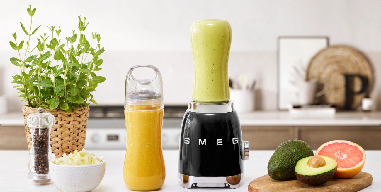 Musta Smeg-tehosekoitin, vaaleanvihreä smoothiekuppi, appelsiininvärinen smoothiepullo, tuore avokado ja greipinpuolikas keittiön tiskillä.