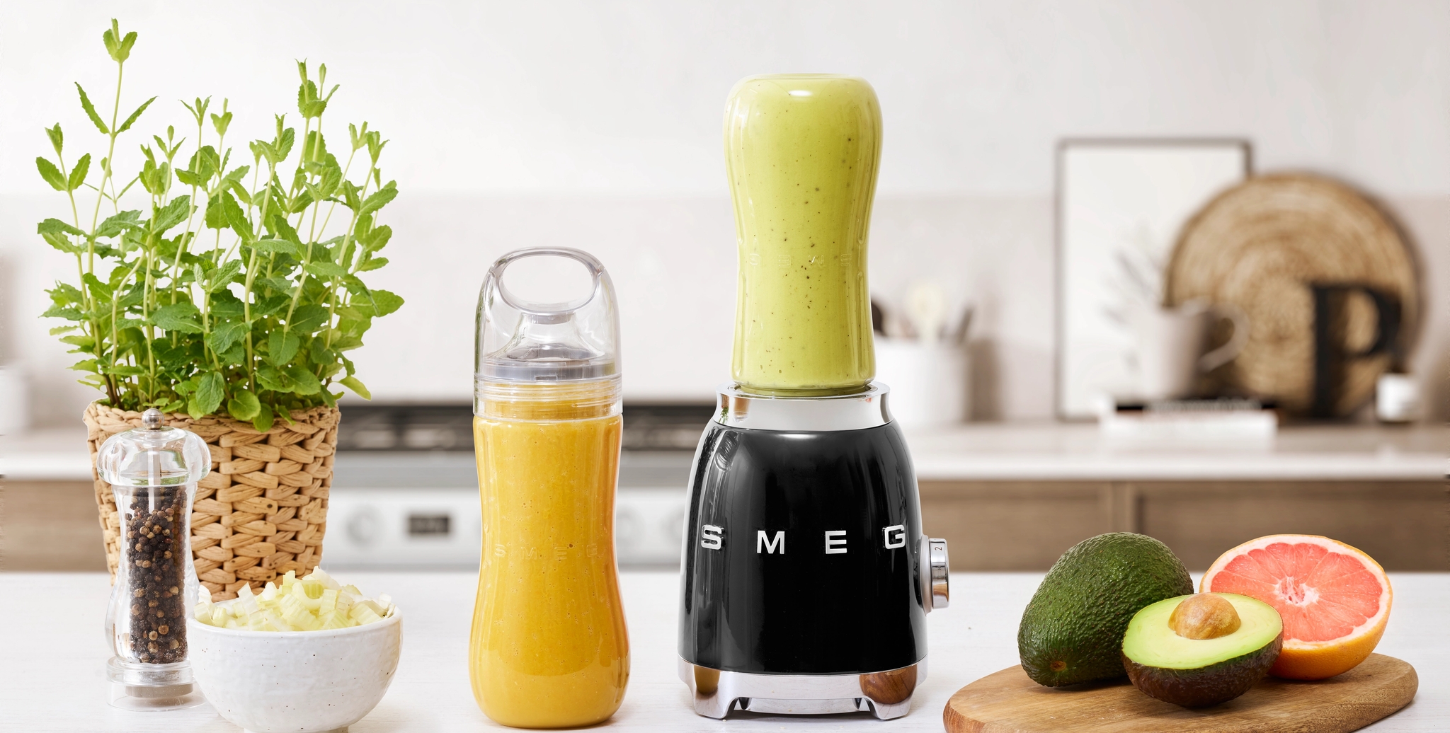 Musta Smeg-tehosekoitin, vaaleanvihreä smoothiekuppi, appelsiininvärinen smoothiepullo, tuore avokado ja greipinpuolikas keittiön tiskillä.
