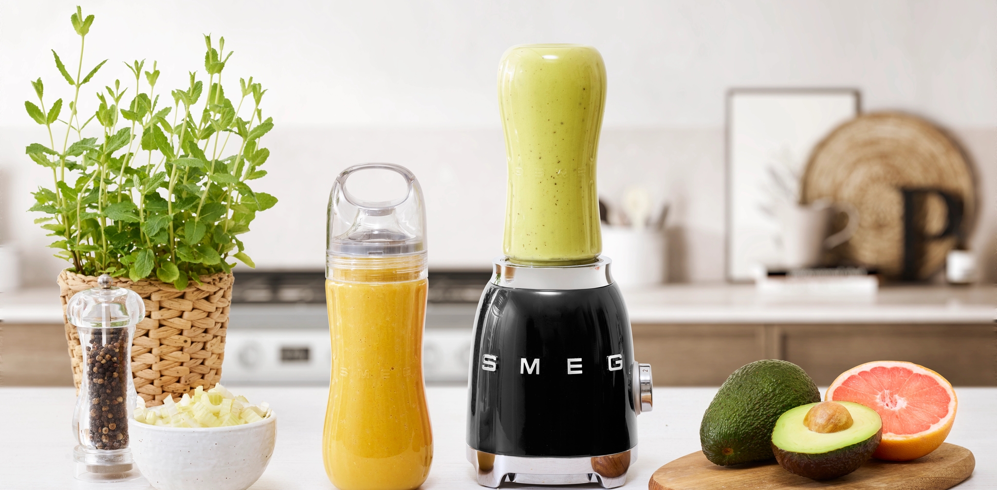 Musta Smeg-tehosekoitin, vaaleanvihreä smoothiekuppi, appelsiininvärinen smoothiepullo, tuore avokado ja greipinpuolikas keittiön tiskillä.