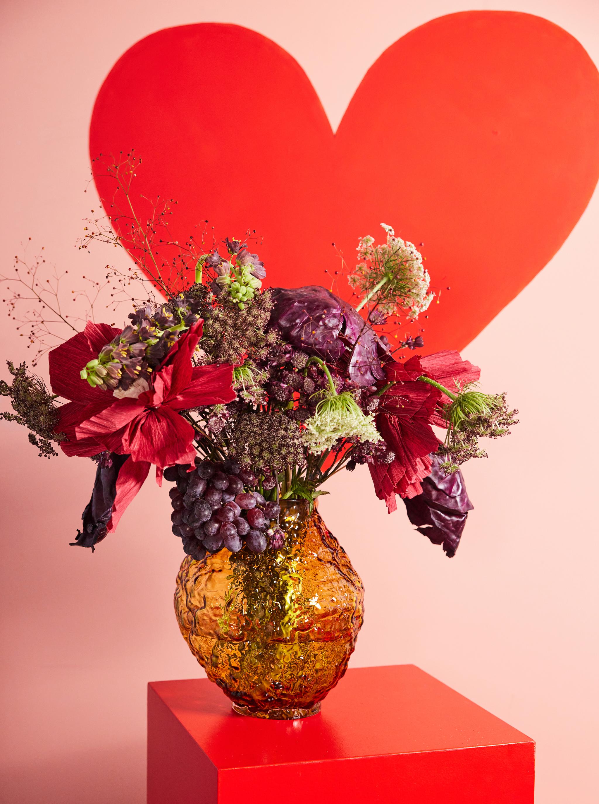 Et slående blomsteroppsett av røde blomster, lilla bladverk og druer i en gullvase på en rød kube, med et stort rødt hjerte i bakgrunnen.