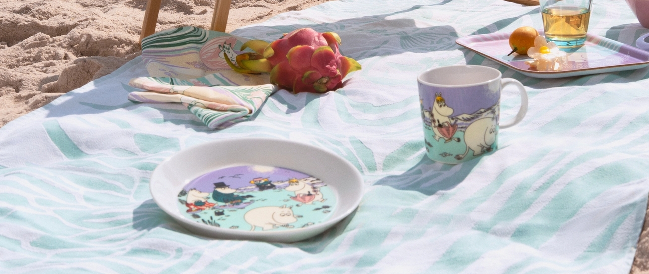 Sommarmuggen 2025, tallriken och brickan i pastelliga färger från Moomin Arabia på en handduk på stranden.