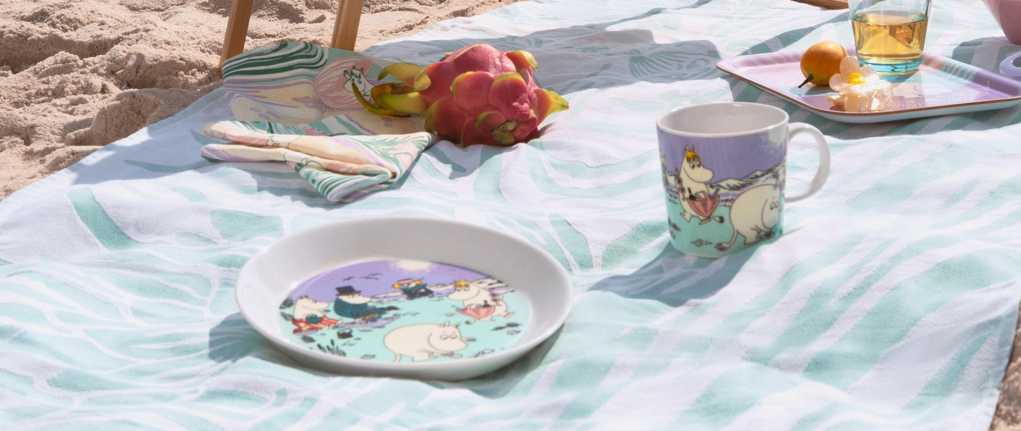 Sommerkoppen 2025, tallerken og brettet i pastell farger fra Moomin Arabia på et håndkle på stranden.