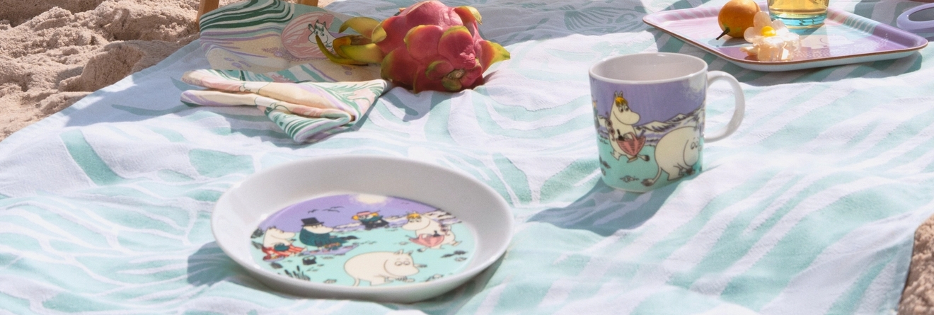 Sommarmuggen 2025, tallriken och brickan i pastelliga färger från Moomin Arabia på en handduk på stranden.