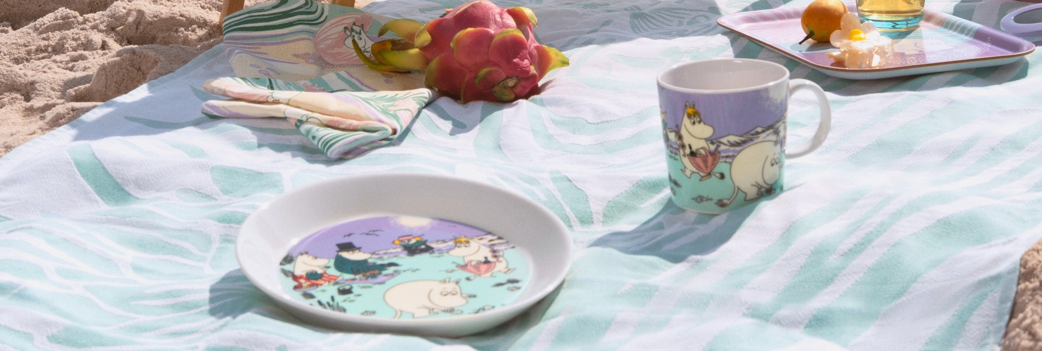 Sommerkoppen 2025, tallerken og brettet i pastell farger fra Moomin Arabia på et håndkle på stranden.