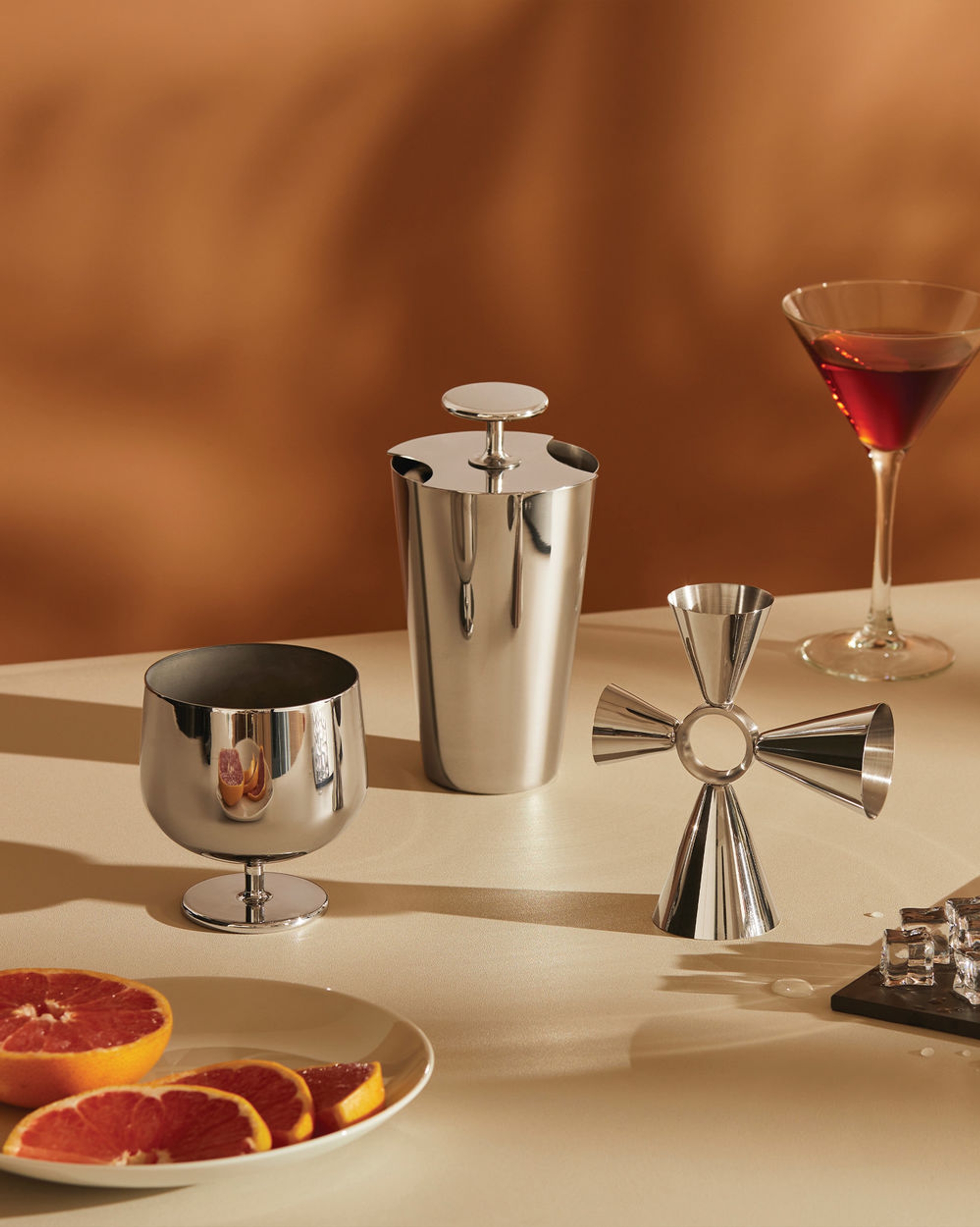 Cocktailverktøy i rustfritt stål, en martini og grapefruktskiver på et bord.