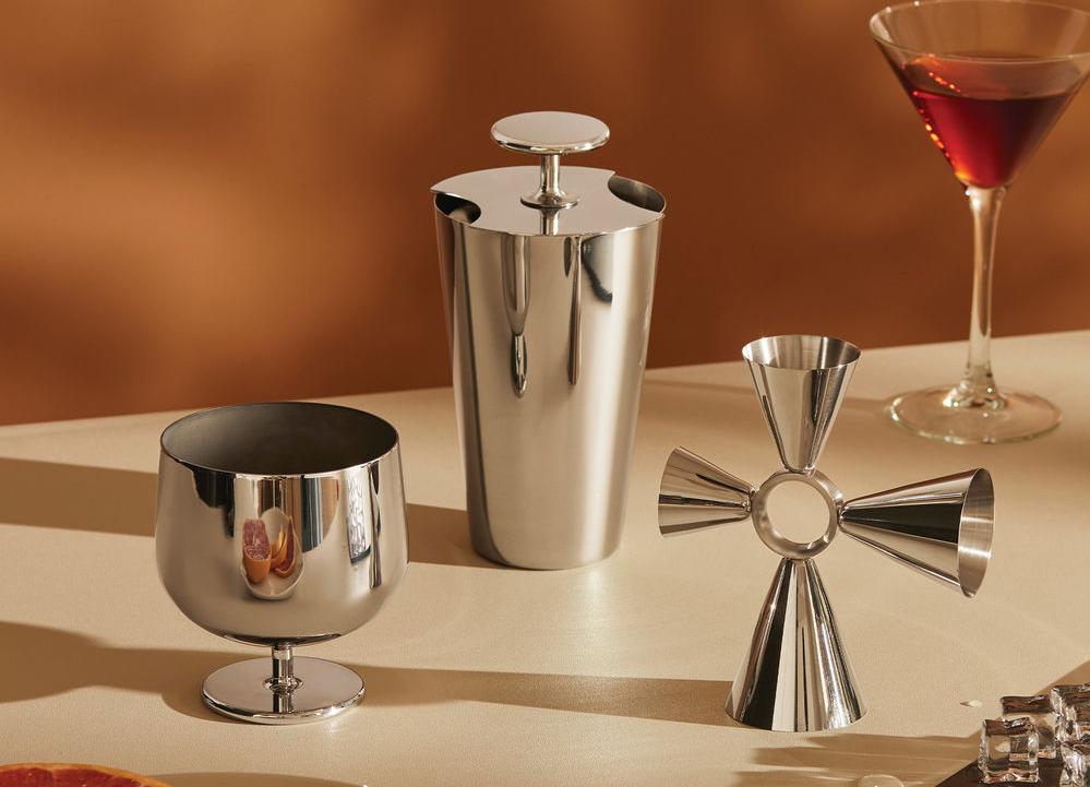 Tyylikäs cocktail-asetelma, jossa on kiiltäviä teräksisiä baaritarvikkeita, martini-lasi ja viipaloitua greippiä lämpimän ruskeaa taustaa vasten.