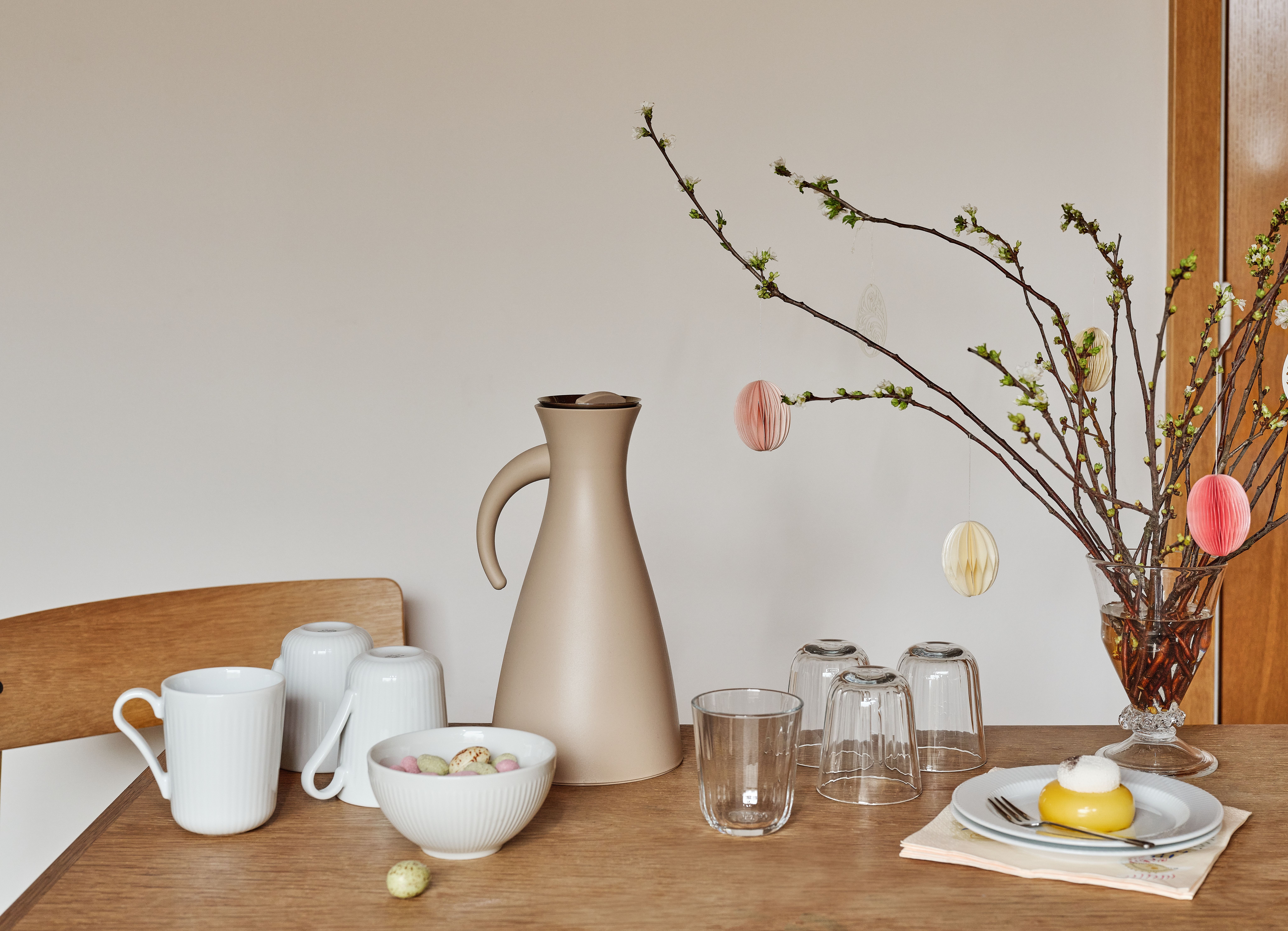 Kevätkattaus, jossa beigenvärinen karahvi, valkoisia mukeja, kulhollinen karkkeja, pieni keltainen kakku ja nupullaan olevia oksia, jotka on koristeltu paperisilla pääsiäismunilla.