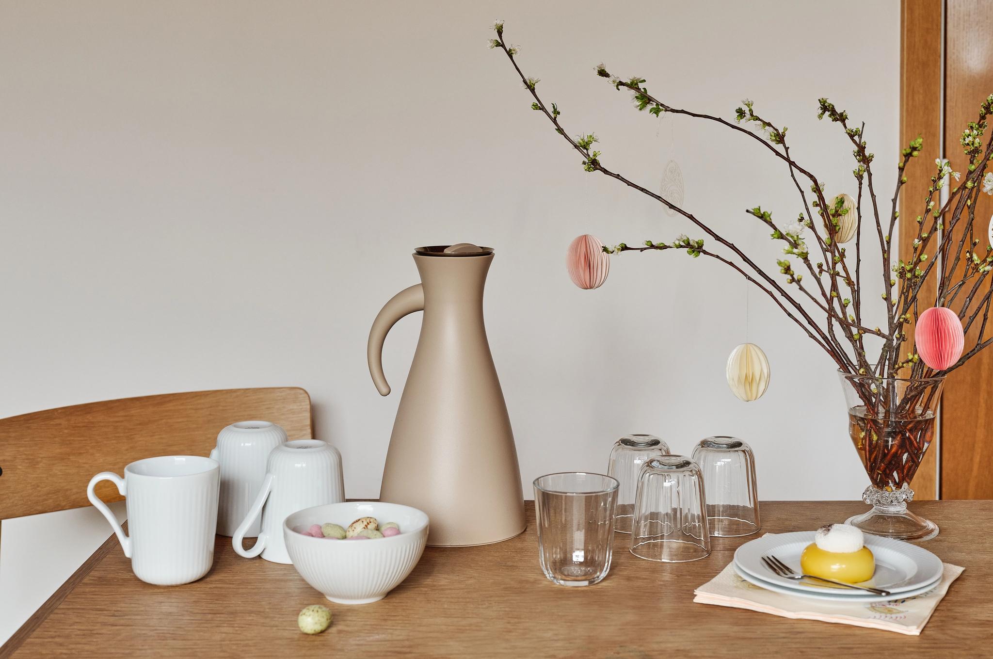 Kevätkattaus, jossa beigenvärinen karahvi, valkoisia mukeja, kulhollinen karkkeja, pieni keltainen kakku ja nupullaan olevia oksia, jotka on koristeltu paperisilla pääsiäismunilla.