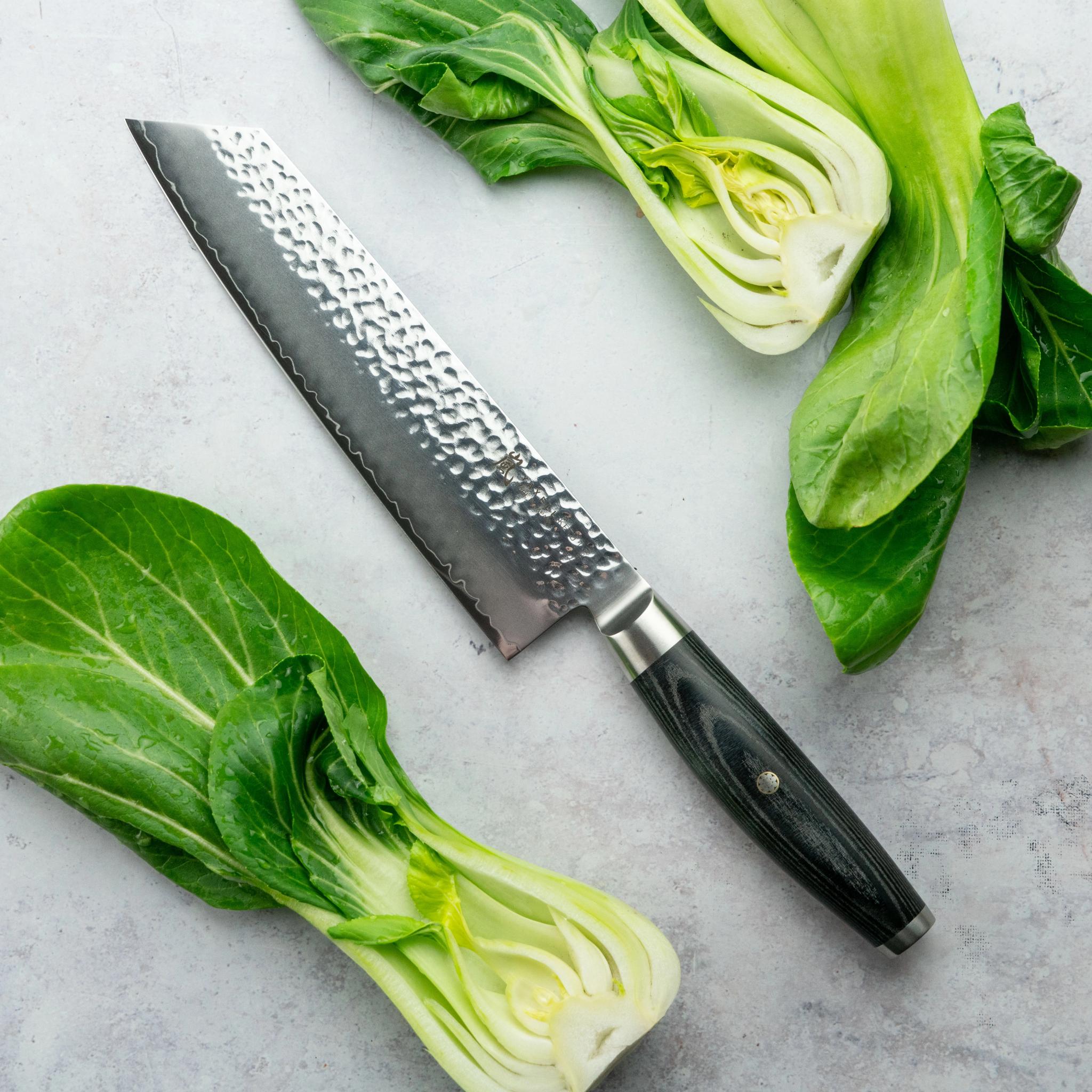 Hamrad kniv bredvid hel och halverad bok choy på en ljusgrå yta.