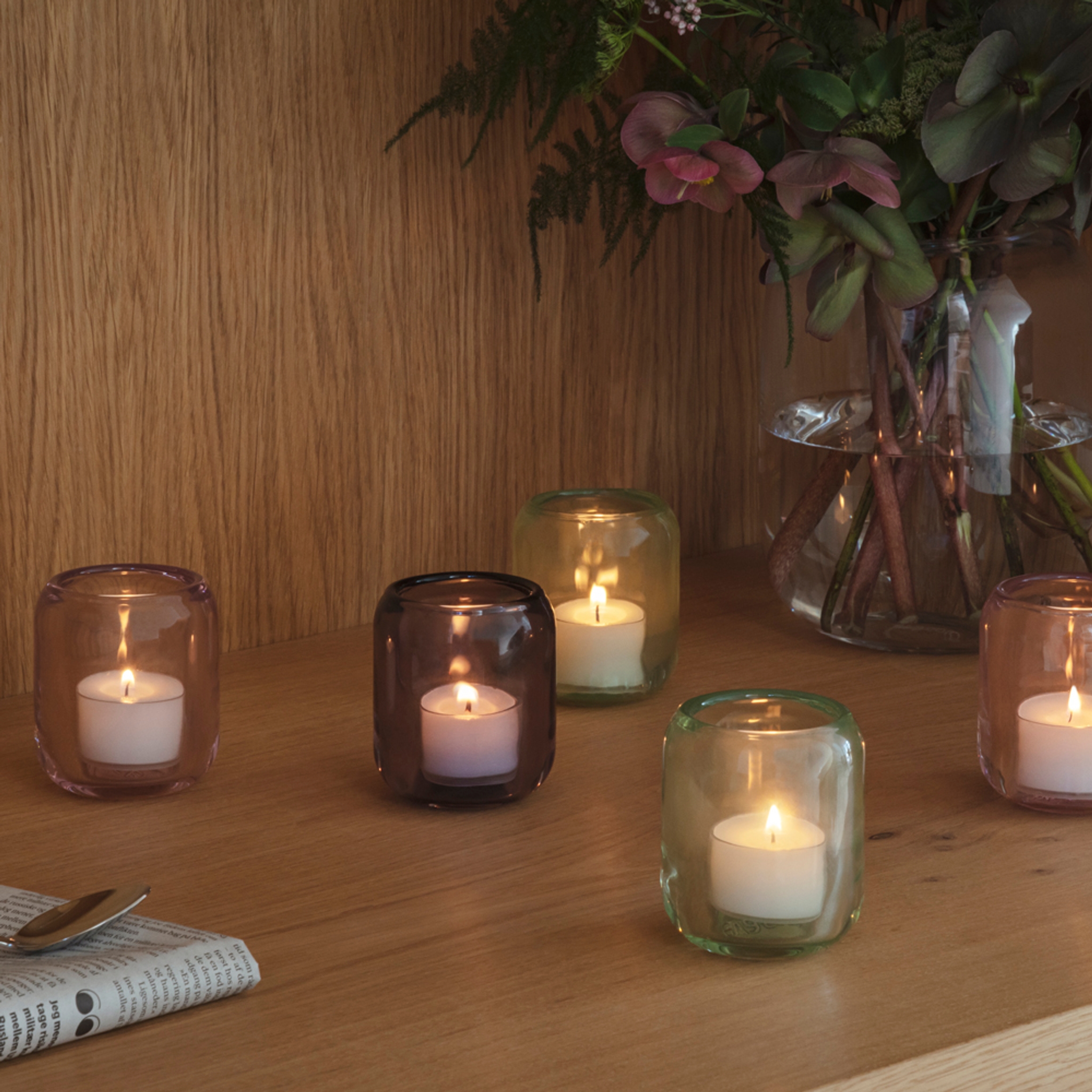 Viisi palavaa teekynttilää värillisissä lasikuvuissa puupinnalla, ja maljakko täynnä kukkia.