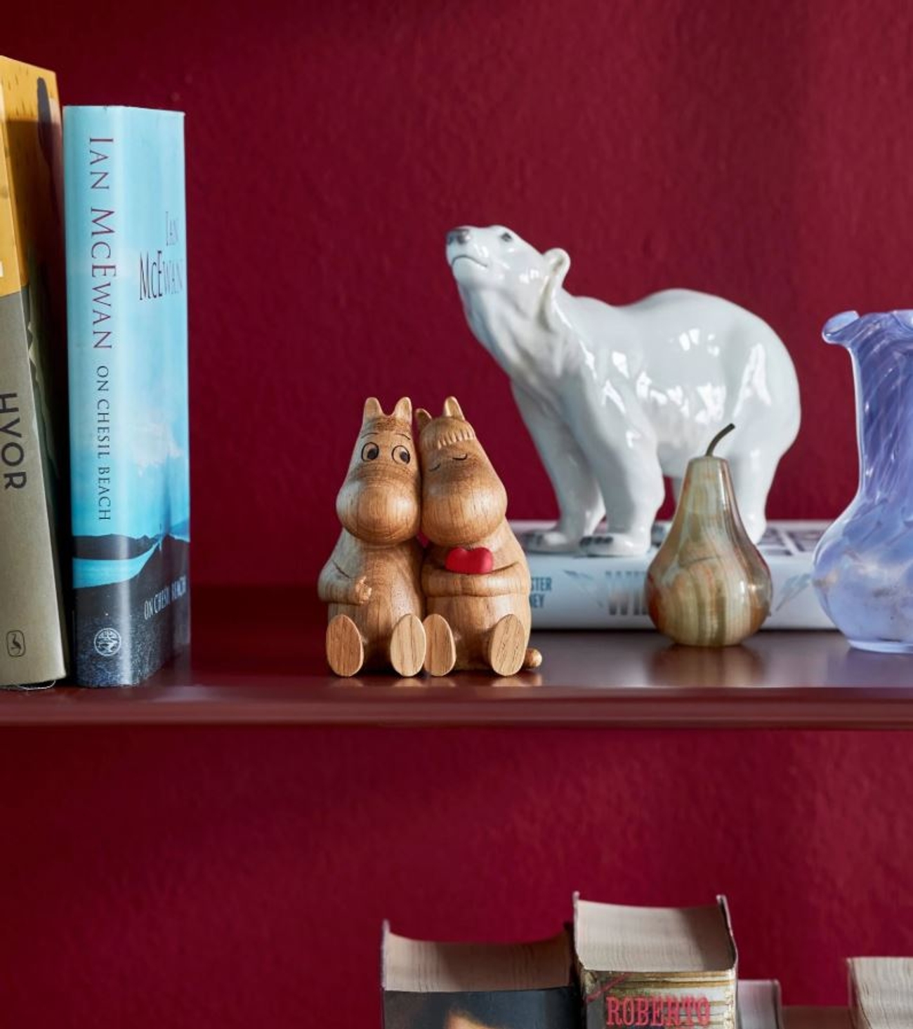 A wooden Moomin couple figurine holding a heart, a white polar bear, and books on a shelf against a red wall.