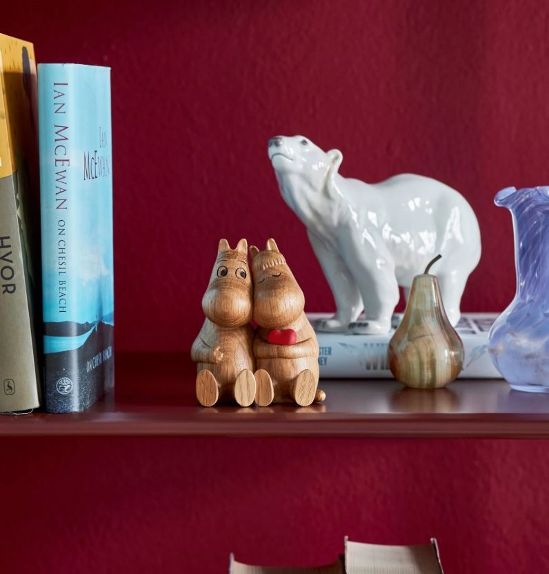 Books, two wooden Moomin figurines embracing with a red heart, a white ceramic polar bear, a pear ornament, and a blue glass vase sit on a shelf against a red wall.