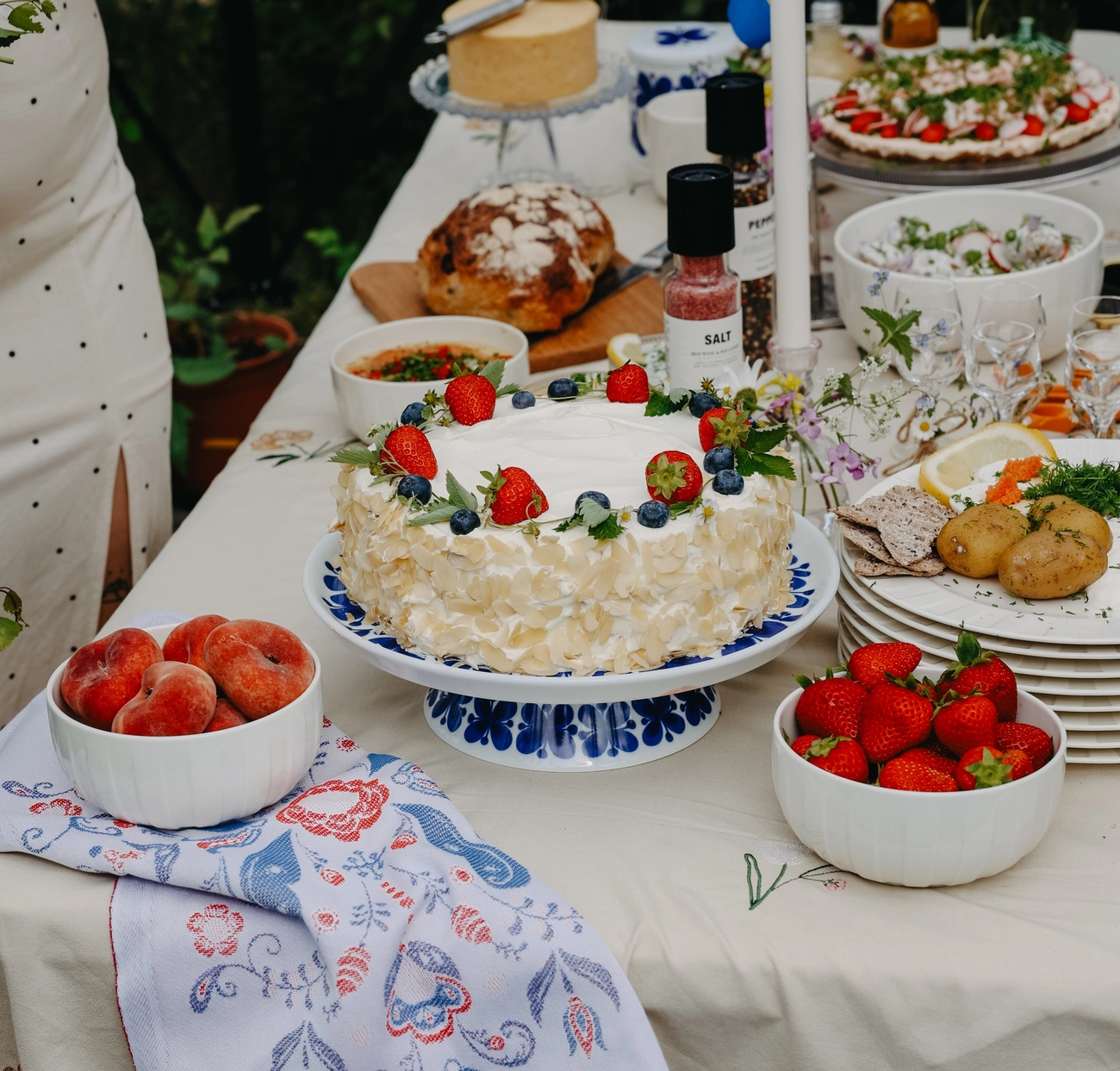Somrig svensk festdukning utomhus med jordgubbstårta, färska bär, potatis och små svenskflaggor på bordet.