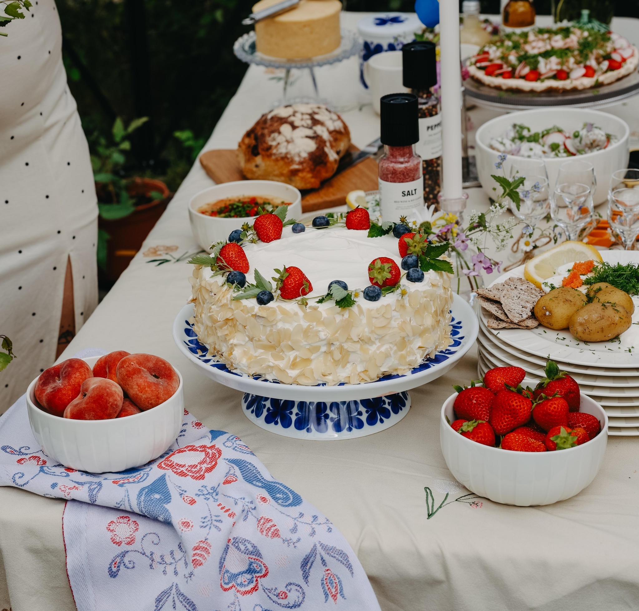 Somrig svensk festdukning utomhus med jordgubbstårta, färska bär, potatis och små svenskflaggor på bordet.
