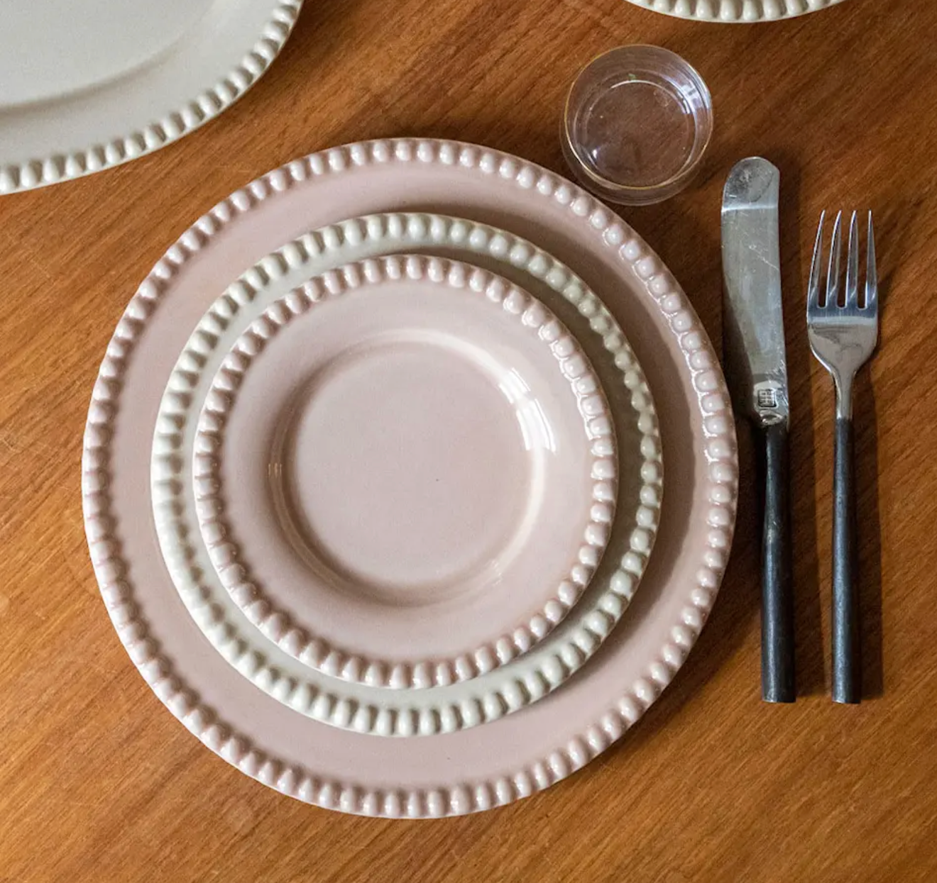 Rosa keramik från PotteryJo Daria – handgjorda tallrikar i olika storlekar med dekorativ pärlkant, dukade på ett träbord med bestick och glas vid sidan.