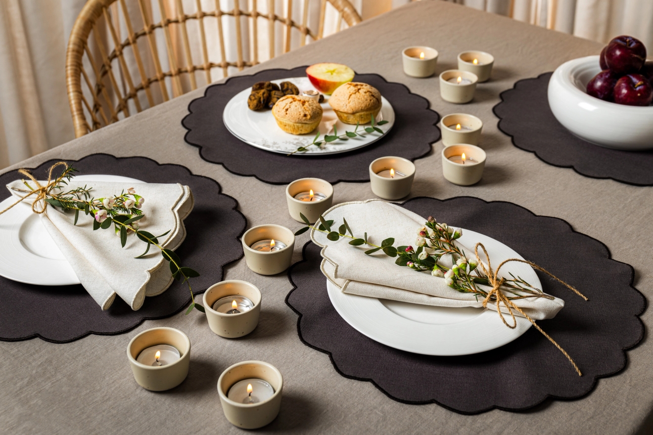 Ett matbord dukat med en ljusgrå duk, mörka vågiga bordsunderlägg, vita tallrikar med benvita servetter dekorerade med grönt och blommor, många tända värmeljus, muffins, bakverk, en äppelskiva och plommon i en skål.