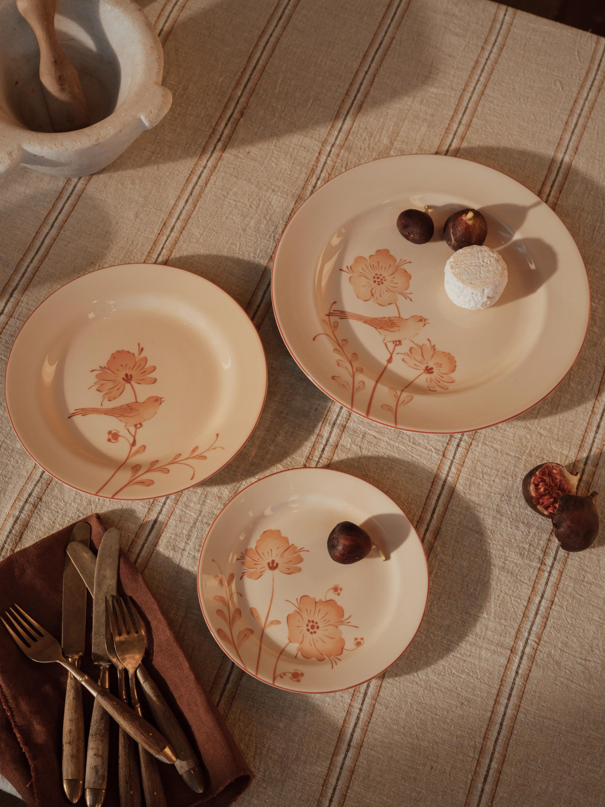 Kremfargede tallerkener med oransje blomster- og fuglemønster, fiken, ost og bestikk på en stripete linduk.