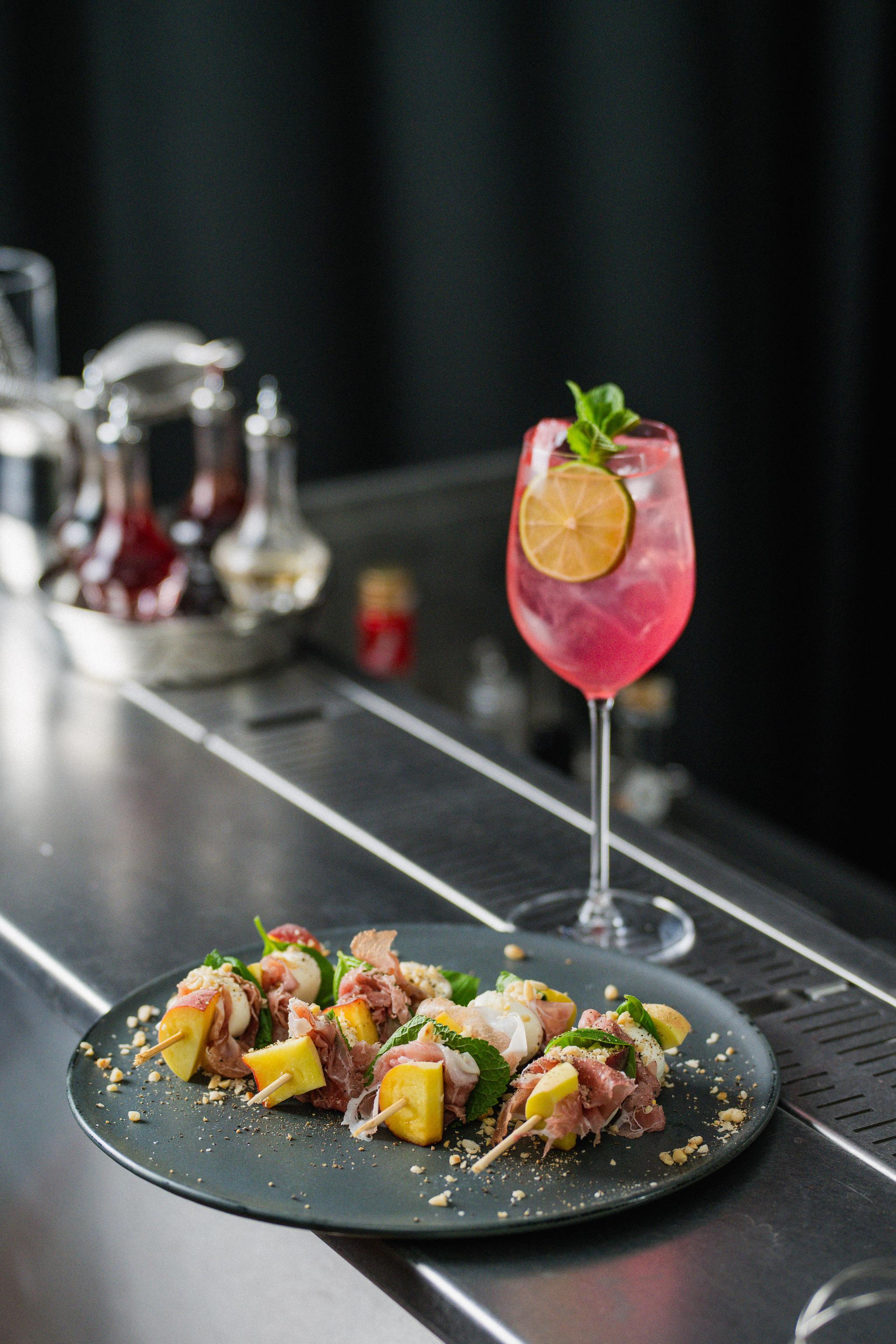 Elegant barservering med färgglad rosa cocktail och tallrik med lyxiga aptitretare på spett.
